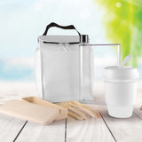 A white picnic pack with a drink bottle, a reusable cup, and eco-friendly utensils, set against a blurred outdoor background.