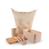 A mesh bag in cream holds a cream mug, wooden cutting board, pink lunch boxes, and pink cutlery.