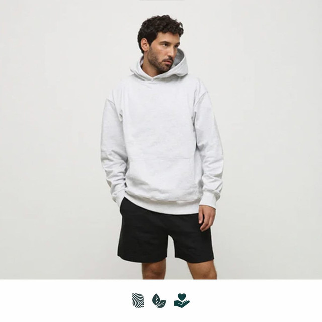 A man wearing a light grey hoodie and black shorts stands against a plain background. The hoodie has a logo.