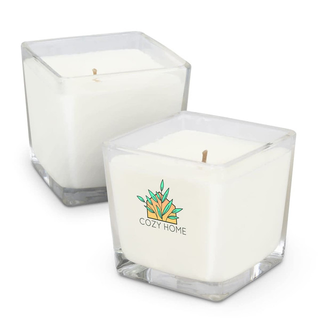 A square, white scented candle in a clear glass container, featuring a logo design. Two candles are shown.