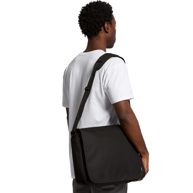 A man with short curly hair is holding a black recycled messenger bag over his shoulder. The bag features a branding logo.