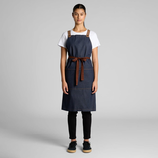 A denim apron featuring brown leather straps and a tie, displayed on a model against a neutral background.
