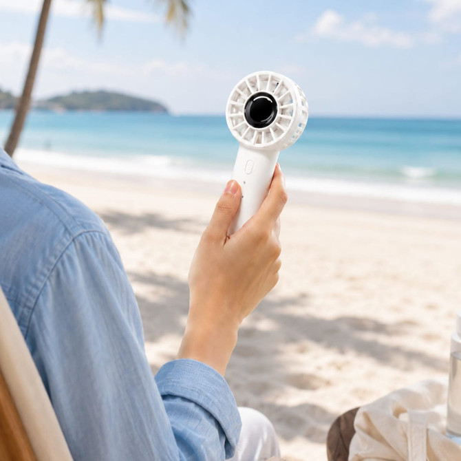 A white rechargeable fan is held by a person, set against a beach background with sand and palm trees.