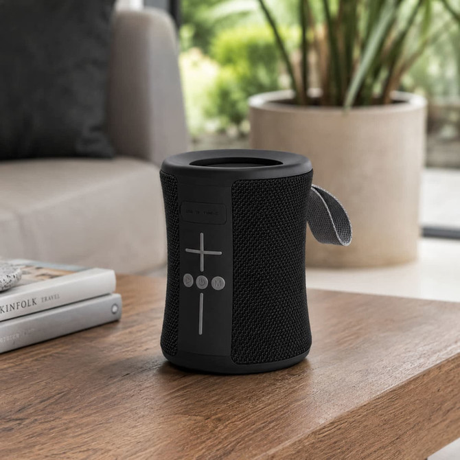 A black portable speaker with buttons on the front, placed on a wooden table beside a book and a plant.