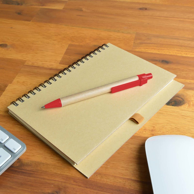 A kraft notebook with a red Matador pen placed on a wooden desk next to a keyboard and a computer mouse.