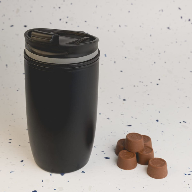 A reusable coffee cup with a sleek black design, featuring a secure lid, accompanied by several chocolate coffee pods.
