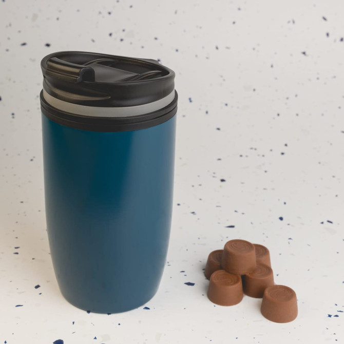 A reusable coffee cup in a teal color with a black lid is positioned beside several chocolate-colored coffee pods.