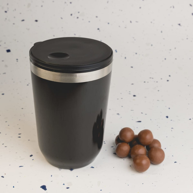 A reusable black coffee cup with a stainless steel band and a black lid, alongside a handful of chocolate balls.