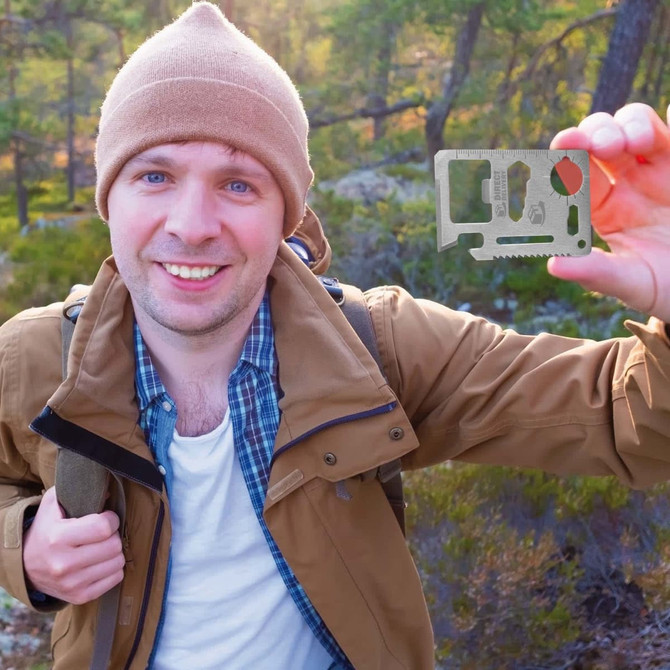 A man in a brown jacket and beige beanie holds up a silver multi-tool in a natural outdoor setting.