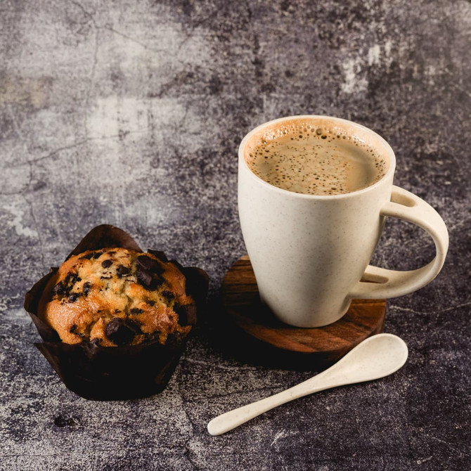A cream-colored reusable coffee cup with a wooden base, spoon, and a chocolate chip muffin. The cup features a logo.