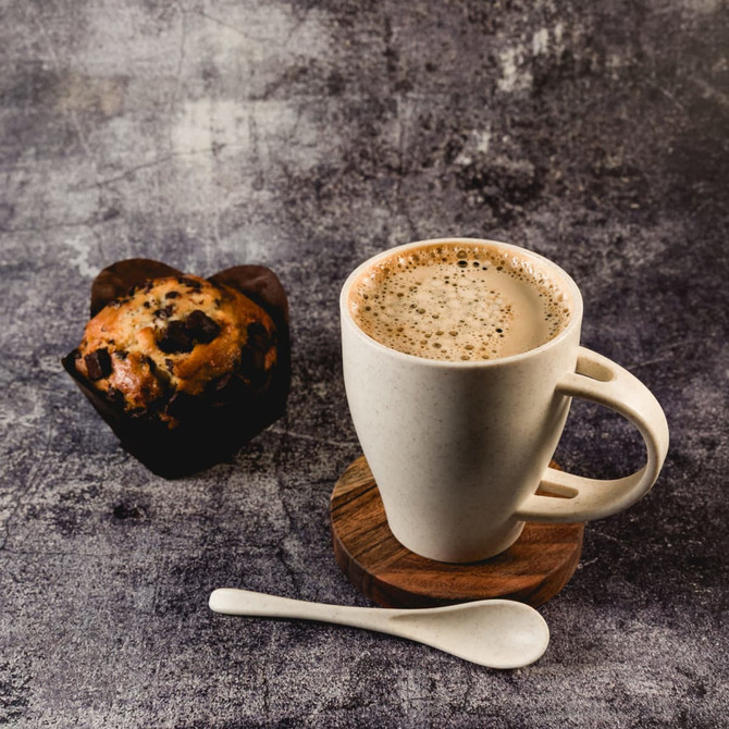 A cream-coloured wheat fibre cup with a spoon, resting on a wooden coaster beside a chocolate muffin.