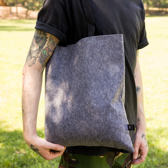 A grey felt tote bag held by a person in a black shirt, featuring a strap and a logo.