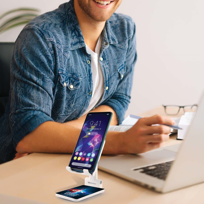 A white phone stand securely holds a mobile device, displayed on a desk with a laptop and a smiling person nearby.