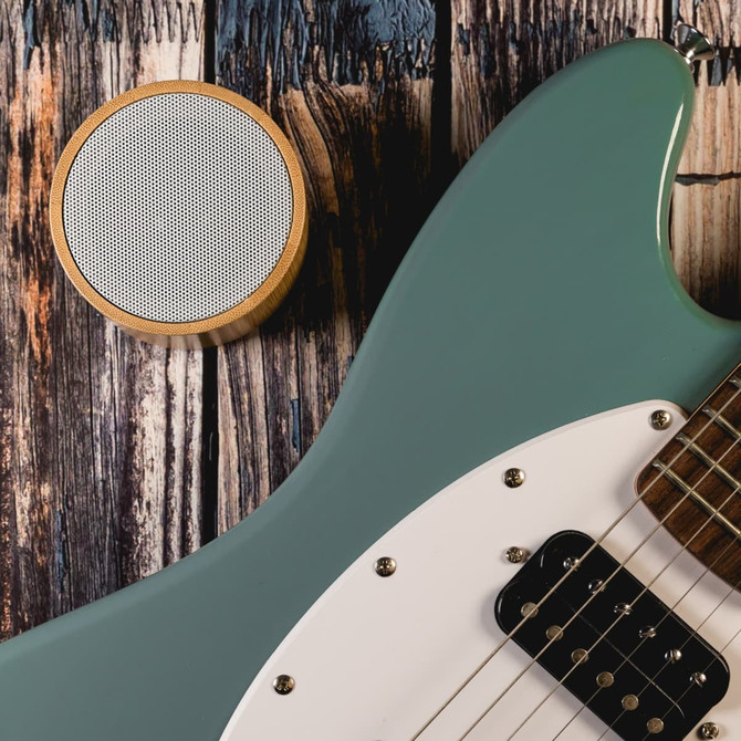 A Bluetooth speaker featuring a bamboo finish is positioned next to a green electric guitar on a wooden surface.