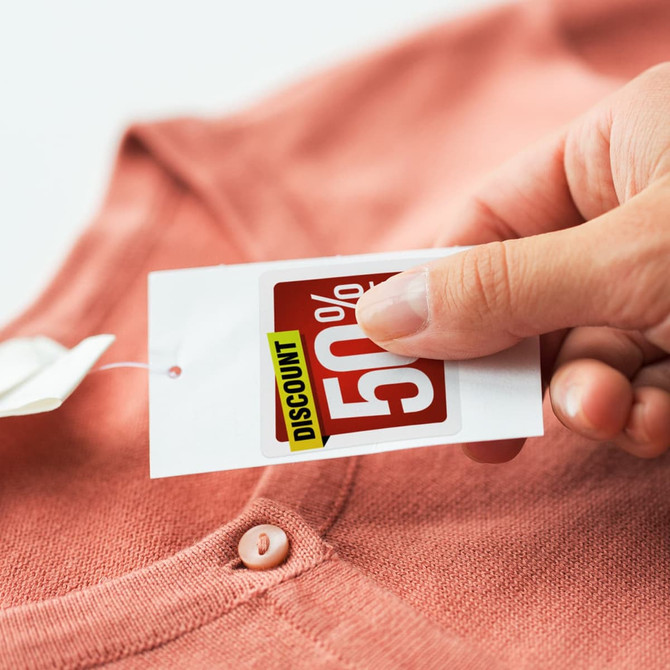 A hand holding a discount tag against a coral-coloured cardigan. The tag displays a percentage discount.