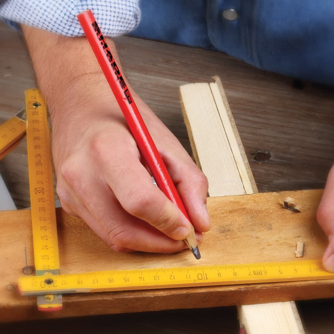 A red carpenter's pencil is held above a wooden surface, marking measurements with a ruler nearby.