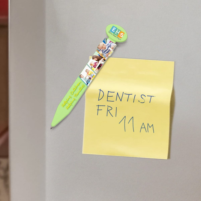 A green plastic pen with a logo, attached to a yellow sticky note on a grey surface.