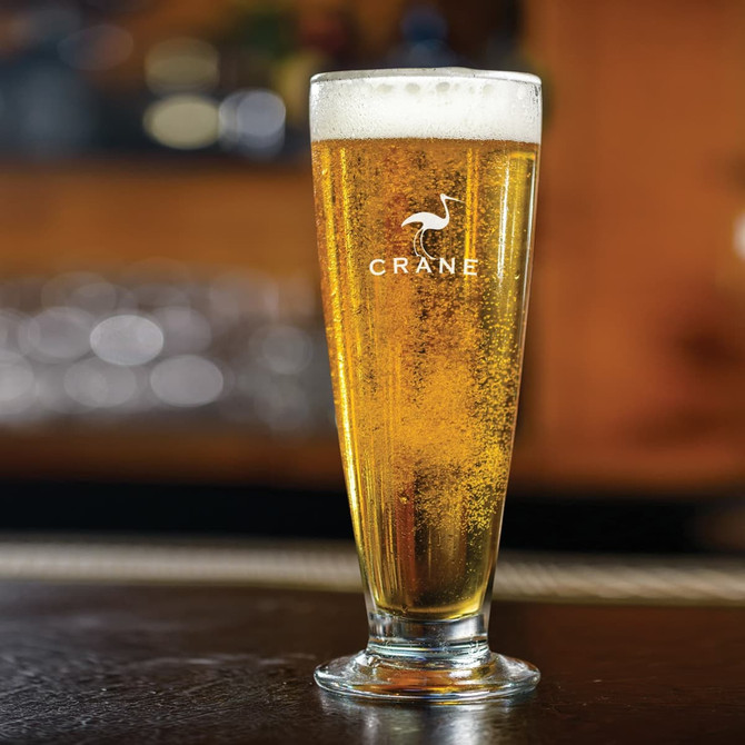 A tall, clear beer glass filled with pale golden beer and a frothy head, featuring a logo.