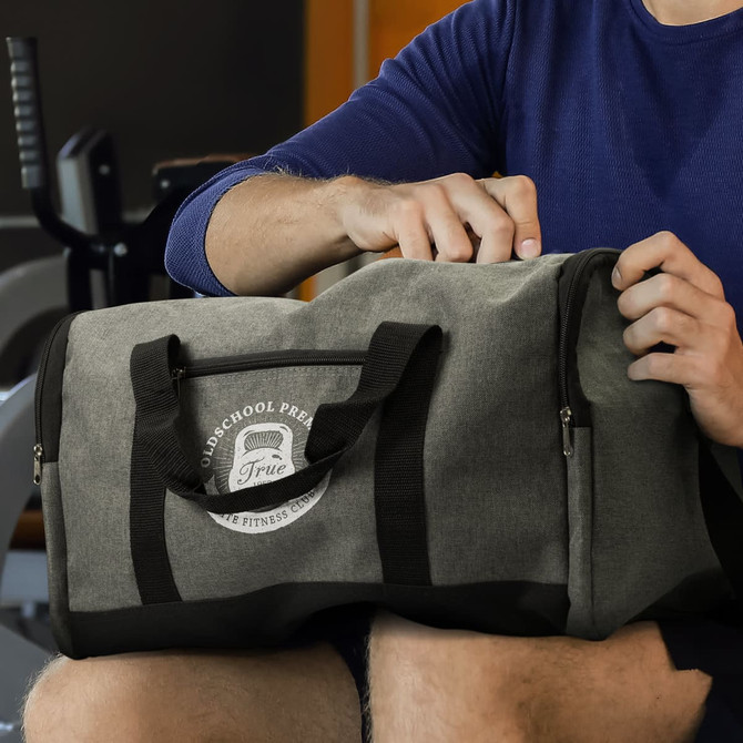 A grey duffle bag with handles and a logo, placed on a person's lap in a gym setting.