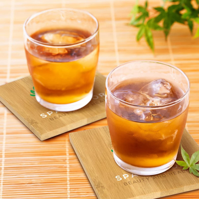 Two clear glasses filled with brown liquid and ice on bamboo coasters, with green leaves in the background.