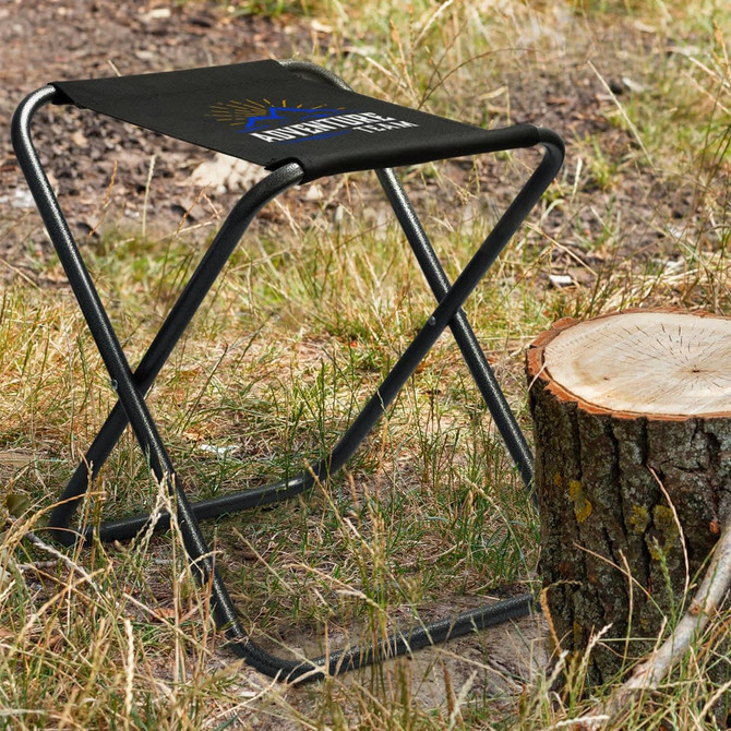 A Quebec Folding Stool with a black seat and a sturdy metal frame, set on grass beside a tree stump.