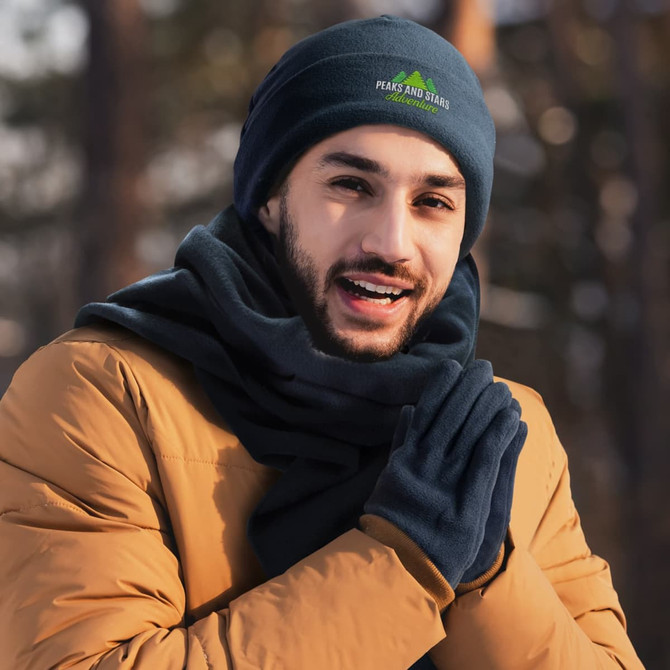 A navy blue fleece beanie and matching gloves set, worn by a smiling person in a warm coat. The beanie has a logo.