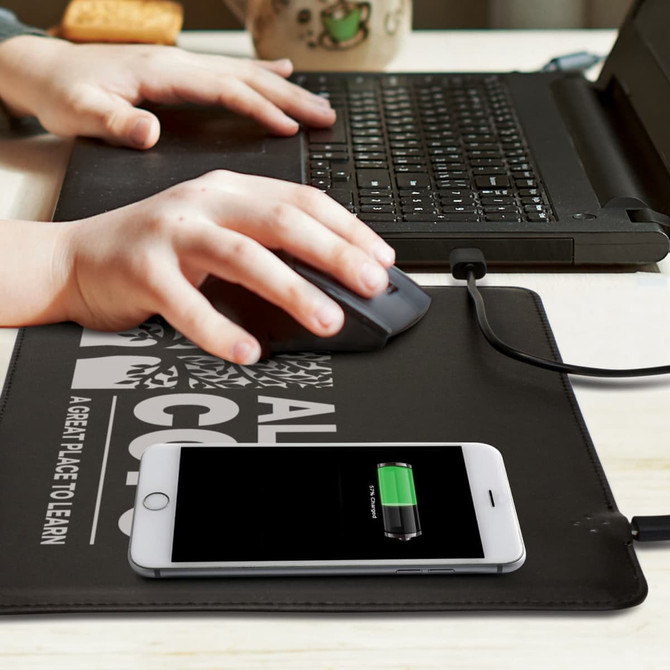 A black wireless charging mouse mat with a phone charging and a laptop in the background, featuring a logo.