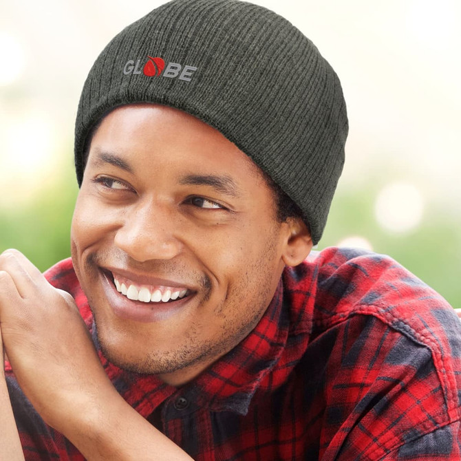 A man smiles while wearing a dark green cable knit beanie with a logo, set against a blurred outdoor background.