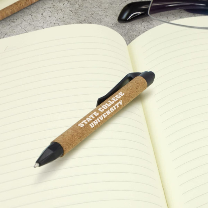 A black plastic pen with a cork grip rests on an open notebook with lined pages. It features a logo.