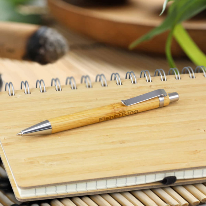 A bamboo pen with a metal clip resting on a wooden surface next to a notebook, featuring a simple design.