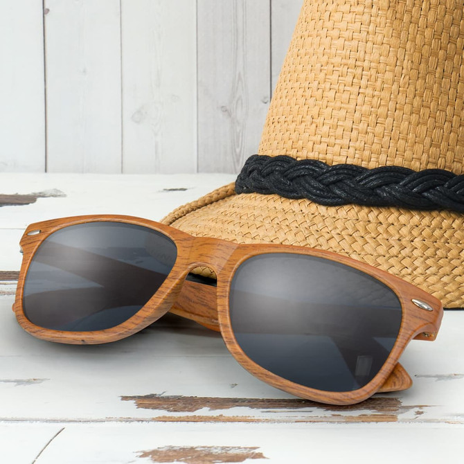 A pair of brown wooden sunglasses with dark lenses, resting on a straw hat with a black band.