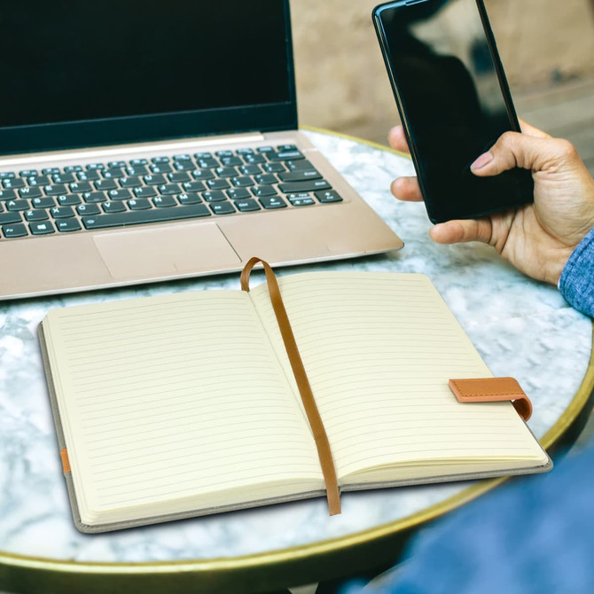 A plain cream notebook with a brown strap, placed on a table next to a laptop and a hand holding a phone.
