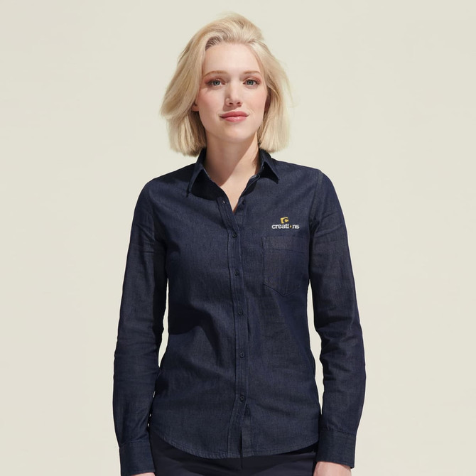 A woman wearing a navy denim button-up shirt with a logo on the chest pocket.