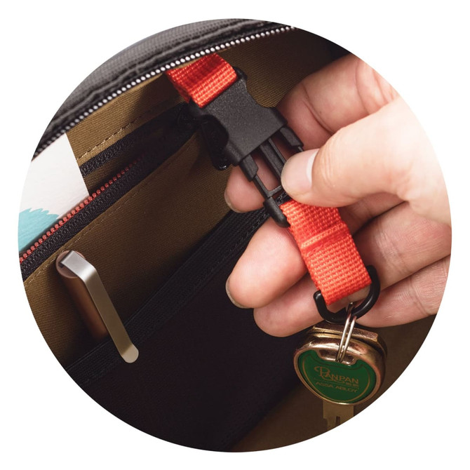 A hand holding a red strap clip inside a black and brown bag, with a keychain and various items visible.