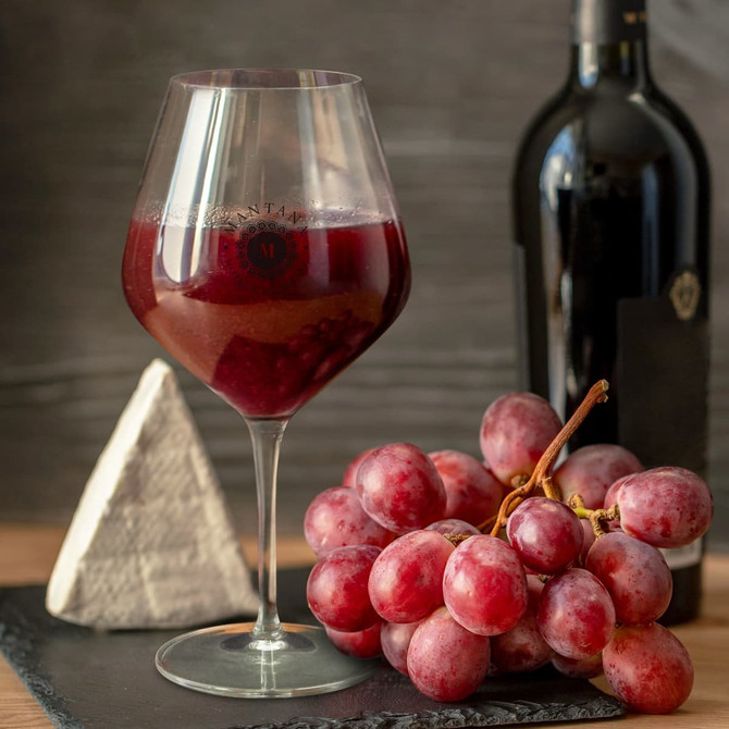 A large, clear wine glass filled with red wine, accompanied by a cluster of red grapes and a bottle of wine.