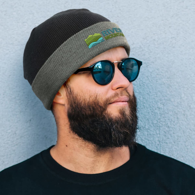 A two-toned beanie in dark brown and grey, worn by a man with sunglasses and a beard, featuring a logo.