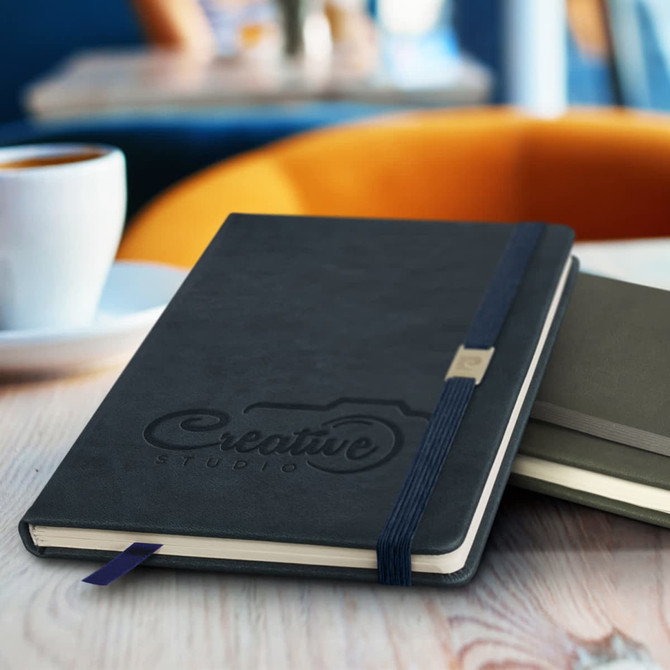 A navy blue notebook with an elastic band and a logo, placed on a wooden table beside a cup of coffee.