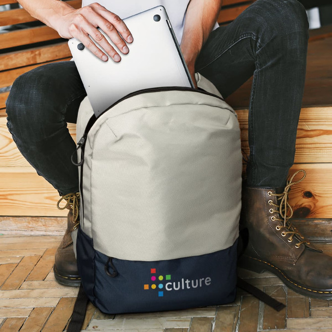 A beige and navy backpack with a logo, held by a person inserting a laptop into it.