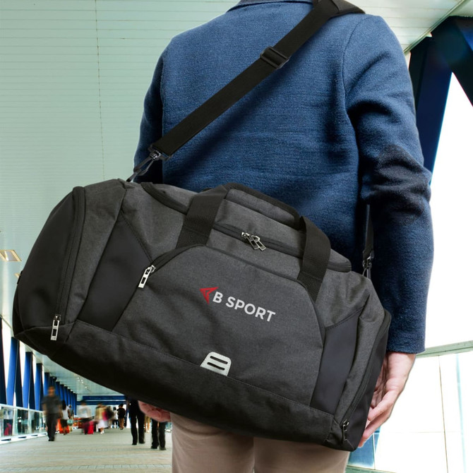 A black duffle bag with grey accents and a logo, carried by a person over their shoulder.