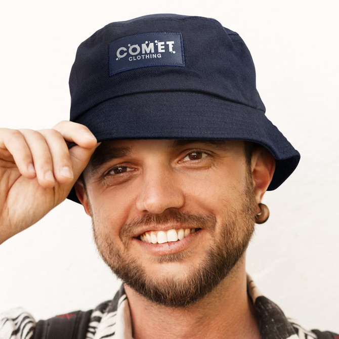 A navy bucket hat with a patch logo is worn by a smiling individual. The hat features a wide brim.