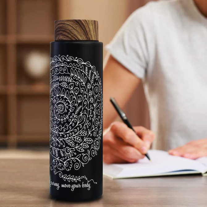 A drink bottle featuring a black exterior with intricate white designs and a wooden lid, positioned beside a notebook.