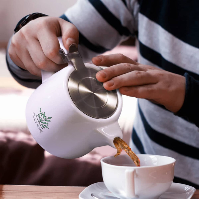 A white teapot with a metal lid, being poured into a cup, held by a person wearing a striped sweater.