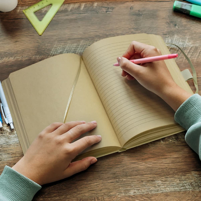 A Kraft notebook open on a desk, with a hand writing in it using a pink pen. Stationery items are nearby.