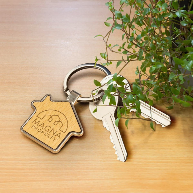 A house-shaped key ring in wood finish, alongside two silver keys and a green plant. It features a brand logo.