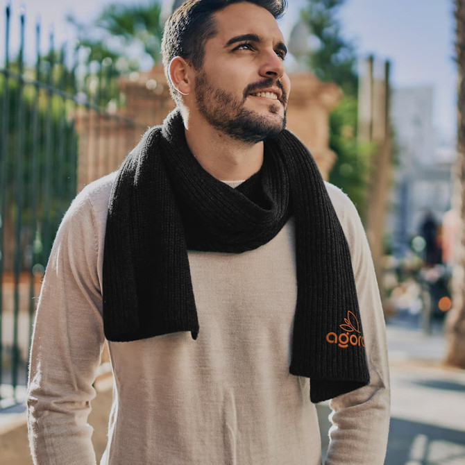 A man wearing a black brushed scarf, standing outdoors with palm trees in the background. The scarf features a logo.