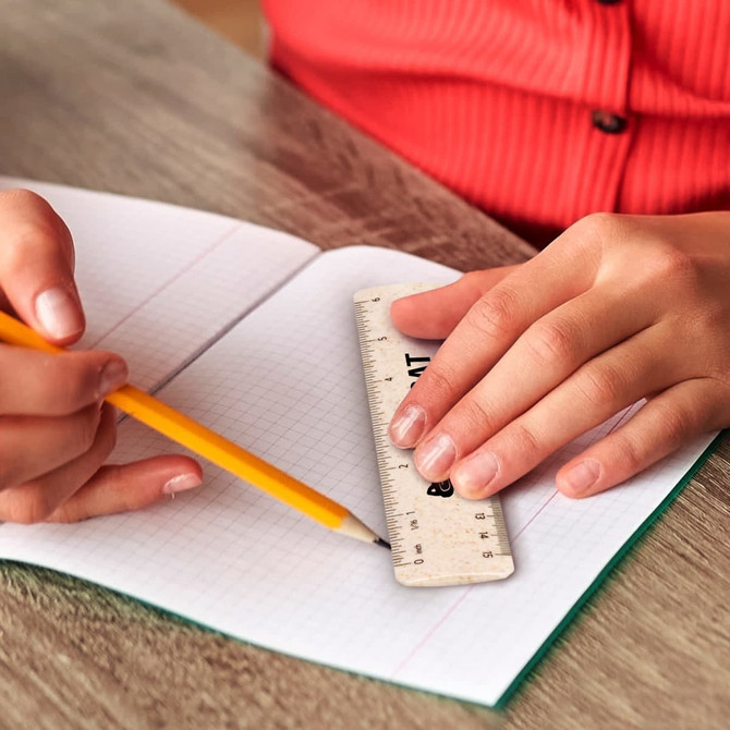 A 15cm ruler rests on a notebook as a hand holds a yellow pencil, on a wooden surface.