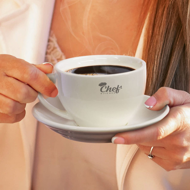 A white chai cup with saucer held by a person, filled with steaming dark liquid, featuring a logo.