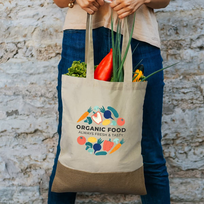 A tote bag in natural colour with a brown base filled with fresh vegetables and a leafy plant. It features a logo.