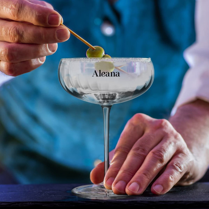 A cocktail glass with a clear rim, filled with a drink and garnished with an olive, held by a hand.