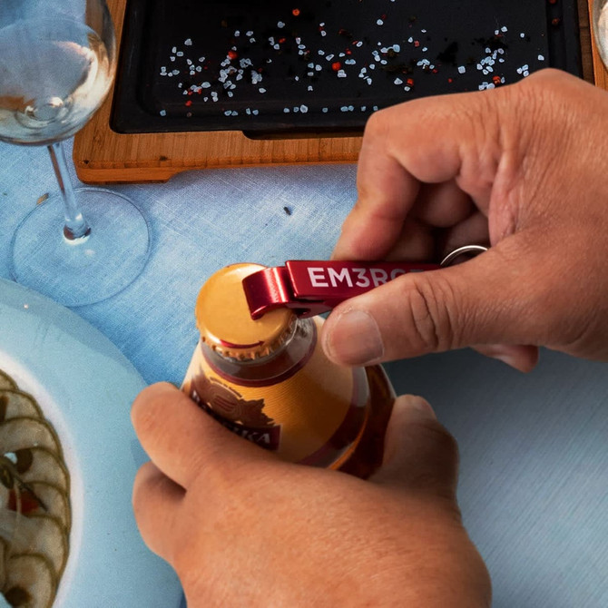 A drink bottle opener in red metal, positioned to open a bottle, surrounded by a table setting with glassware and plates.